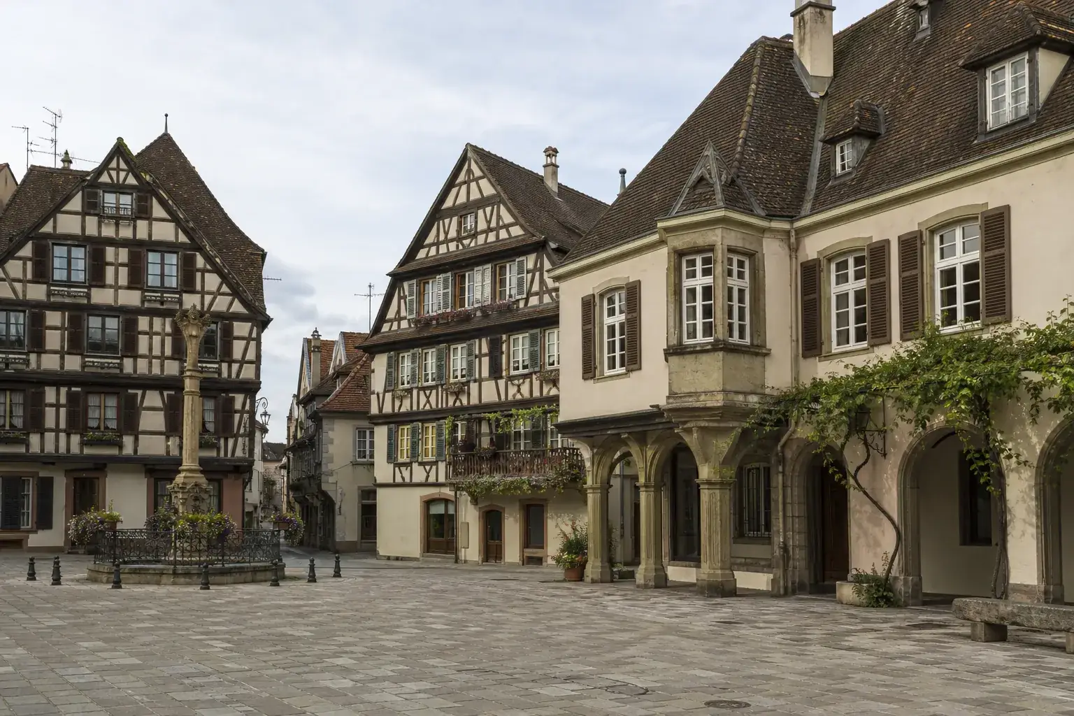 Place pavée d’un centre-ville alsacien cernée de façades à colombages, lumière douce de fin de matinée